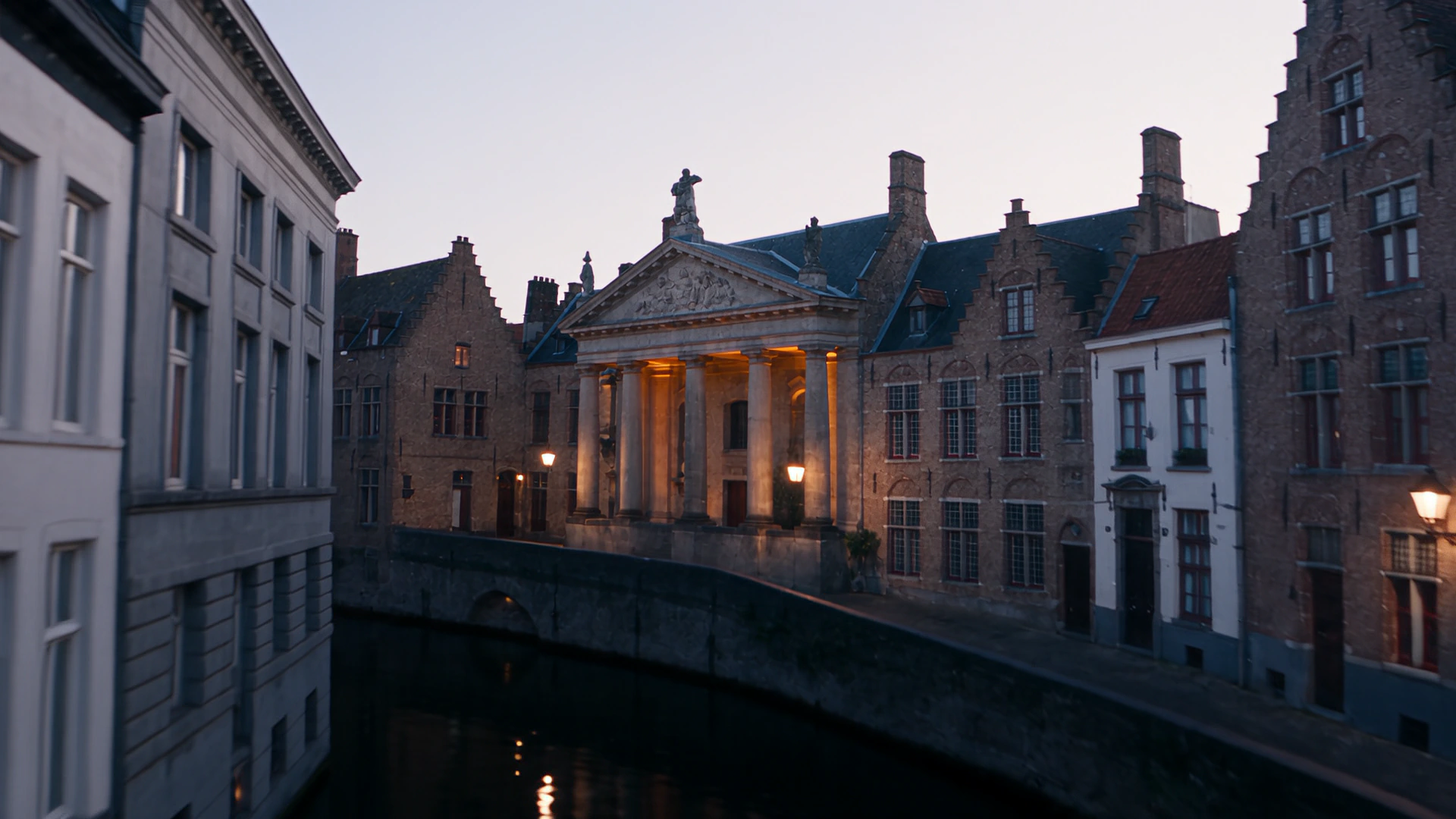 A canal in Bruges at dusk with buildings standing on both banks and a lit portico on the far side.